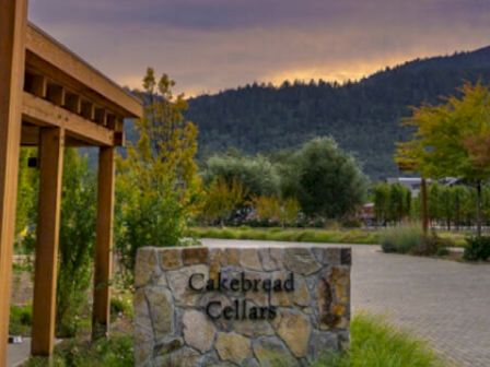 The image shows a stone sign that reads "Cakebread Cellars" with a scenic background of trees and hills under a cloudy sky.
