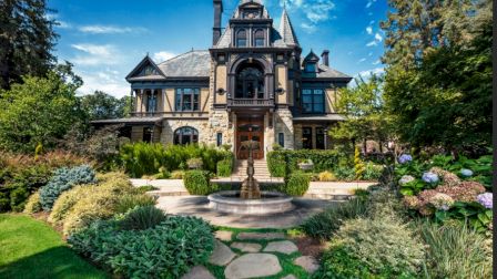 A large, ornate Victorian-style mansion with a lush garden and stone pathway, featuring a central fountain in the foreground.