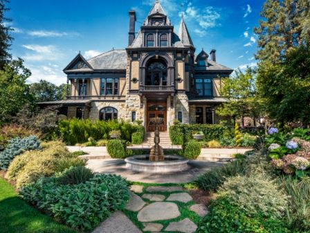 A large, ornate Victorian-style mansion with a lush garden and stone pathway, featuring a central fountain in the foreground.