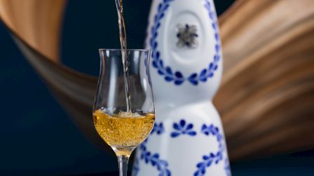 A decorative bottle with blue patterns is in the background, while a golden liquid is being poured into a clear glass in the foreground.