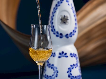 A decorative bottle with blue patterns is in the background, while a golden liquid is being poured into a clear glass in the foreground.
