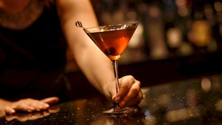 A person holding a martini glass with a cocktail and an olive, set against a dimly lit bar background.