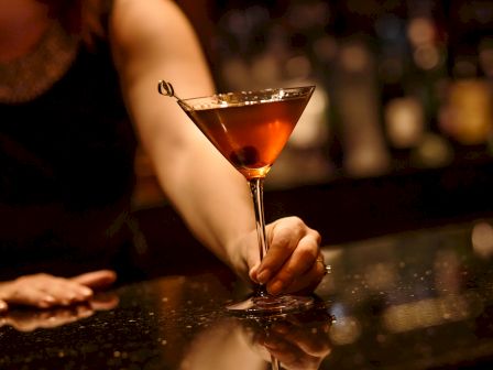 A person holding a martini glass with a cocktail and an olive, set against a dimly lit bar background.