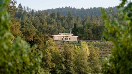 A house is nestled in a lush, forested landscape with trees and hills in the background.