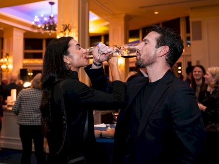 A man and woman are at an indoor event, linking arms and drinking from glasses with people mingling in the background.