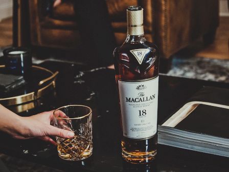 A bottle of Macallan 18-year-old whisky and a glass being held by a hand are on a table.