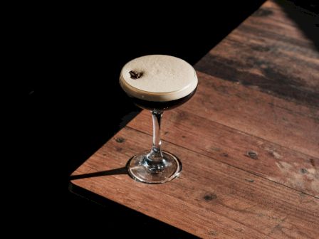 A cocktail in a coupe glass with a coffee bean garnish sits on a wooden table, lit dramatically by sunlight.