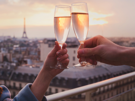 Two people clink champagne flutes in a sunset cityscape celebration, with the Eiffel Tower visible in the distance.