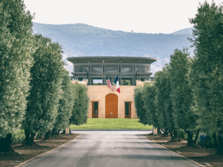 A building with a curved driveway, two flags, surrounded by trees, and hills in the background, under a cloudy sky.