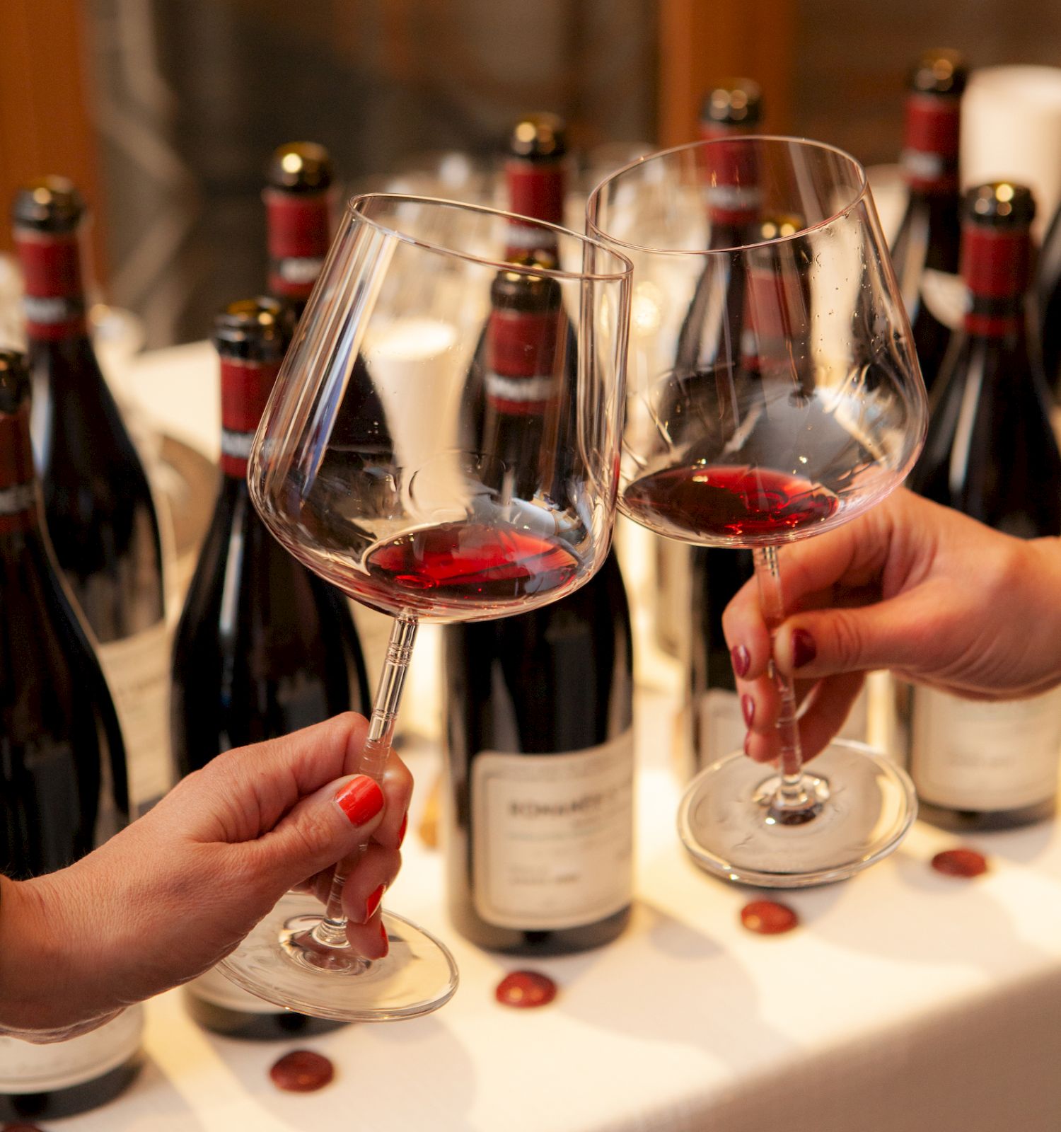 Two people clink glasses of red wine in front of several bottles on a table, celebrating or toasting.