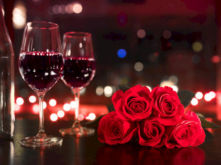 Two glasses of red wine and a bouquet of red roses are set on a table with a bokeh effect in the background, creating a romantic ambiance.