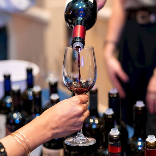Pouring wine into a glass at a tasting, with bottles lined up in the background, as two hands share the pour.
