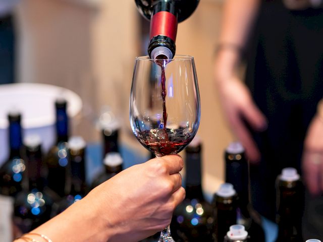 Pouring wine into a glass at a tasting, with bottles lined up in the background, as two hands share the pour.