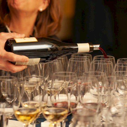 A person pours red wine from a bottle into many wine glasses on a table, with others watching in the background.