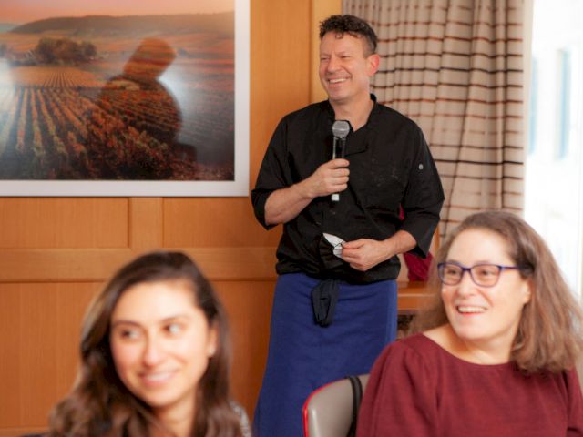 A smiling man in a black shirt and apron speaks into a microphone at a small event, while two amused women sit nearby in a cozy room.