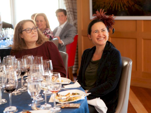 Two women sit at a table with multiple wine glasses and plates; one smiles warmly while others chat in the background at a formal meal.