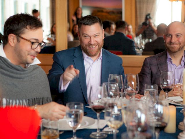 Three men at a restaurant table share a lively moment, with wine glasses, candles, and a bar-style backdrop in the background.