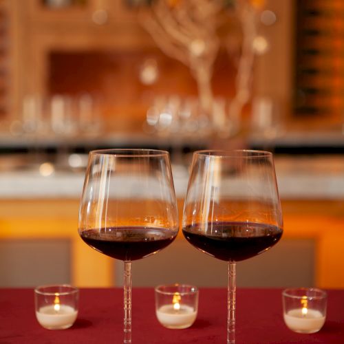Two glasses of red wine sit on a red tablecloth beside lit candles, with a softly blurred toast backdrop.