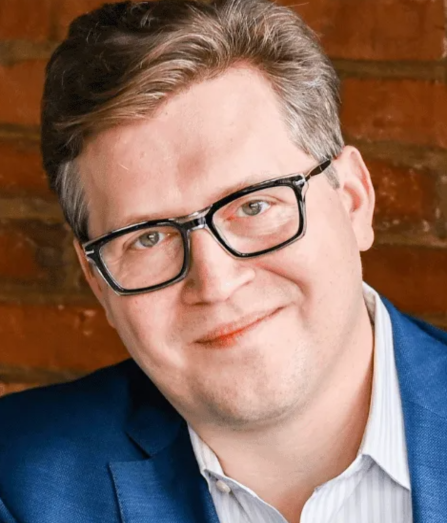 A man wearing black glasses and a blue blazer smiles at the camera against a brick wall background.