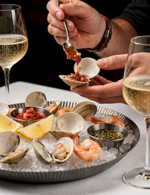 A chef serves a shellfish morsel with red sauce from a spoon onto a small shell at a chilled seafood platter with glasses of wine.