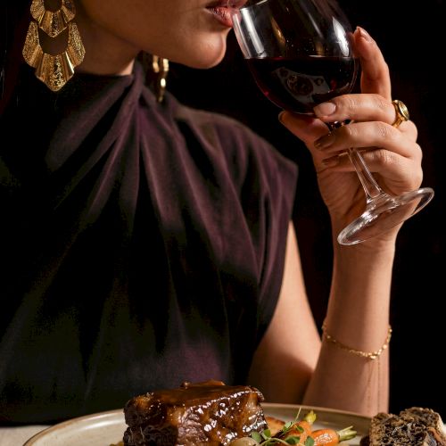 A glamorous woman in a party dress sips red wine while seated at a plated dinner with steak and vegetables.