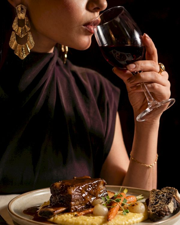 A glamorous woman in a party dress sips red wine while seated at a plated dinner with steak and vegetables.