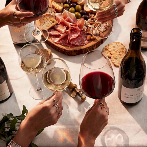 Friends clink red wine glasses over a shared charcuterie board of cured meats, bread, olives, and grapes.