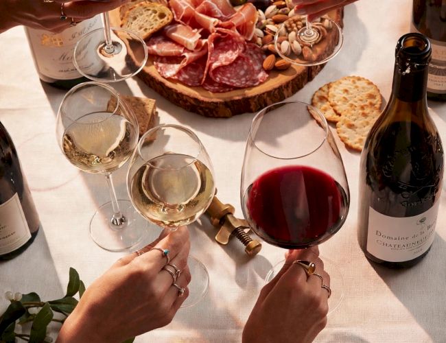 Friends clink red wine glasses over a shared charcuterie board of cured meats, bread, olives, and grapes.