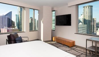 A modern hotel room with large windows, a bed, a wall-mounted TV, and cityscape views. There's a chair and a small side table.