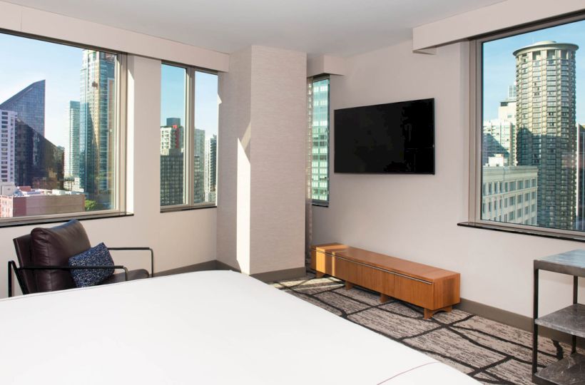 A modern hotel room with large windows, a bed, a wall-mounted TV, and cityscape views. There's a chair and a small side table.