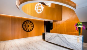 A modern lobby area with wooden walls, a large clock, marble reception desk, and decorative lighting and flowers on the desk.