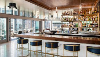 A modern bar with high stools, a well-stocked shelf of bottles, and a view of city buildings through large windows.