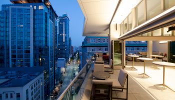 A modern rooftop terrace with seating and tables overlooks a city street lined with tall, illuminated buildings under a dusky blue sky.