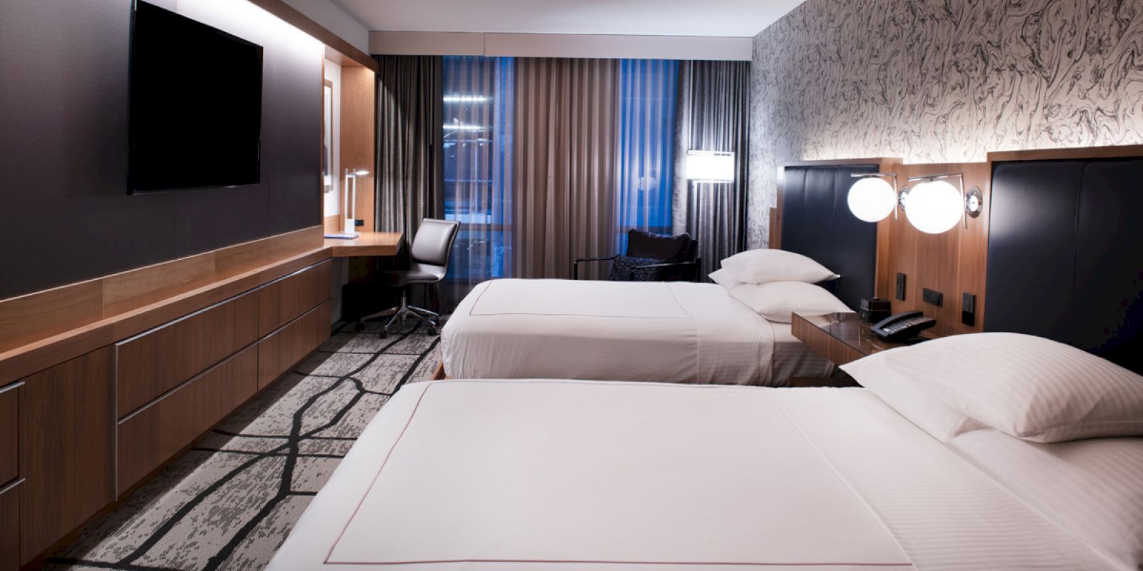 The image shows a modern hotel room with two beds, a TV, a desk, and large windows with curtains. The decor is sleek and minimalistic.