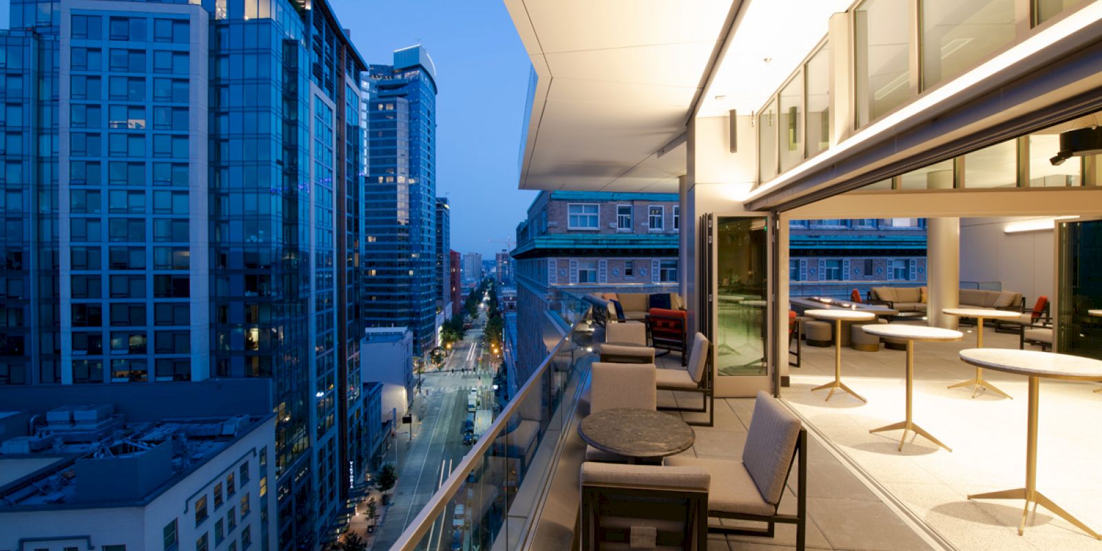 A modern balcony overlooks an urban street at dusk, with tall buildings and lights illuminating the city scene.
