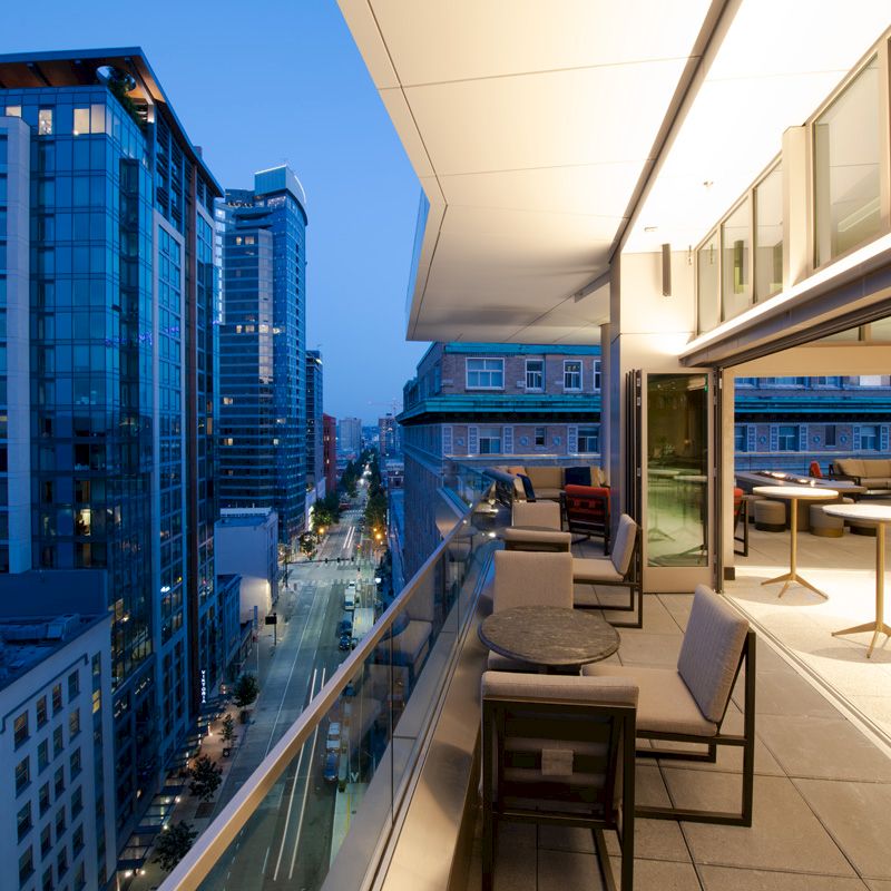 A spacious balcony overlooks a cityscape at dusk, featuring outdoor seating and tables, modern buildings, and illuminated windows.