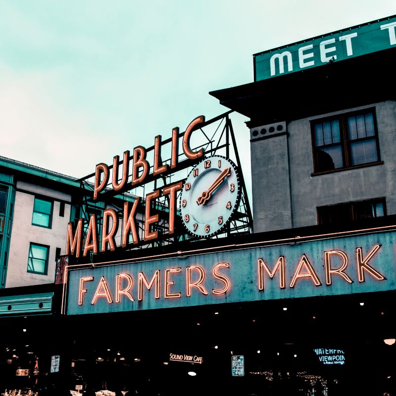 The image shows a vintage neon sign for a public farmers market on a building with a clock displaying the time, set against a cloudy sky.
