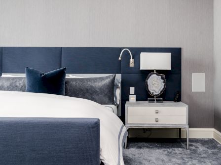 The image shows a modern bedroom with a stylish bed, blue headboard, pillows, and a white nightstand with a lamp and decor item.