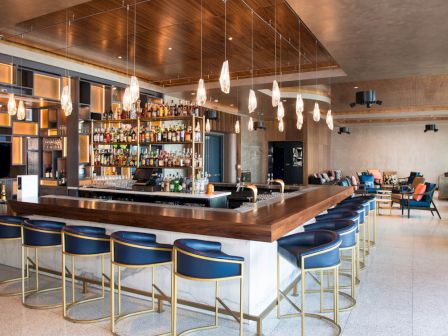 A stylish bar with a wooden counter, blue cushioned bar stools, and hanging pendant lights. Shelves stocked with bottles and seating area.