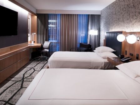 A modern hotel room with two beds, a desk, and a TV. The decor is sleek with patterned wallpaper and contemporary lighting fixtures.