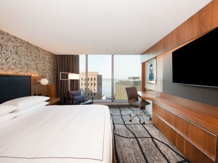 A modern hotel room with a large bed, desk, and chair, facing a window with a city view. A flat-screen TV is mounted on the wall.