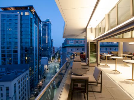 A modern cityscape at dusk featuring tall buildings and a stylish balcony with seating and tables overlooking the street below.