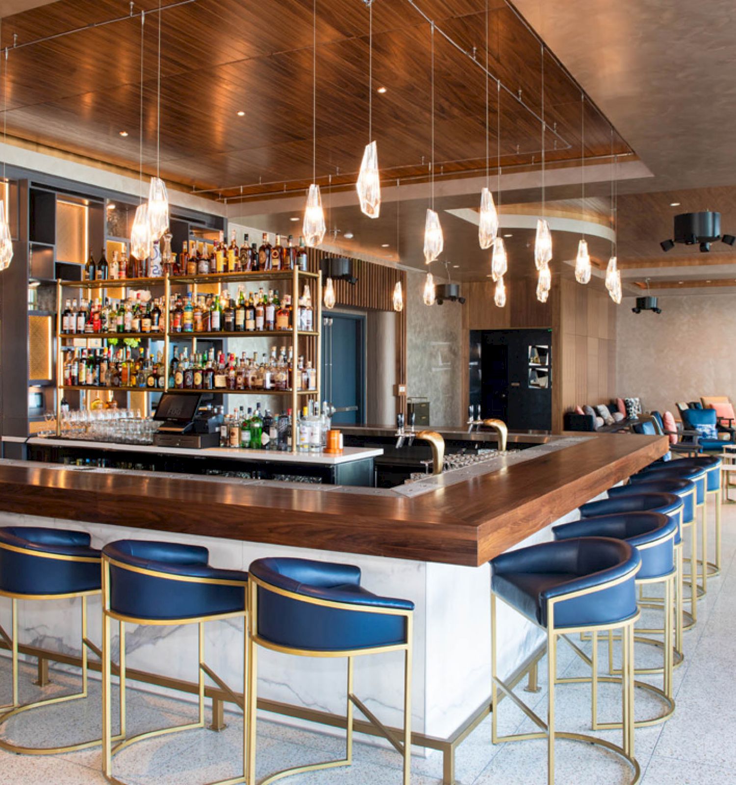 A modern bar with a wooden counter, blue stools, and hanging lights. Shelves of bottles and a seating area are in the background.