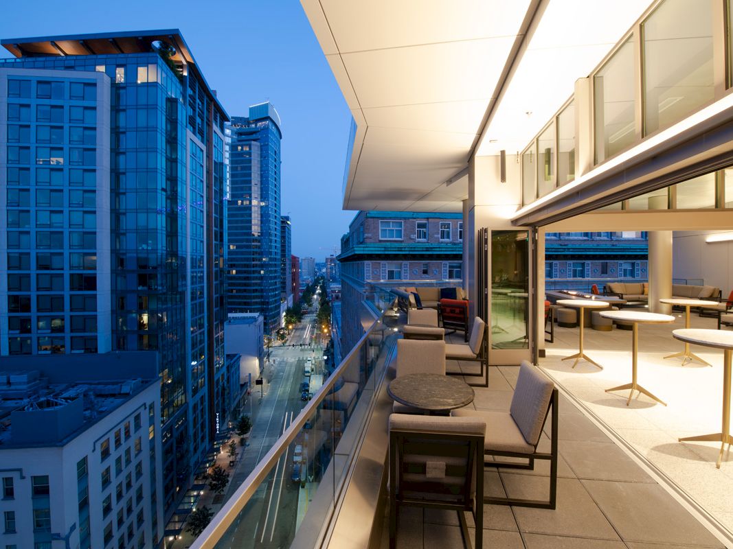 A modern cityscape at dusk, featuring a rooftop terrace with tables and chairs overlooking illuminated skyscrapers and a street below.