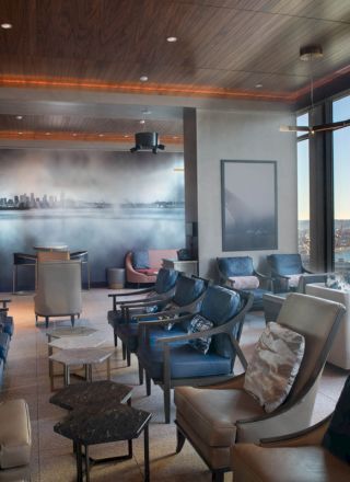 The image shows a modern lounge with comfortable chairs, a cityscape mural, panoramic windows, and a view of a ferris wheel and waterfront.
