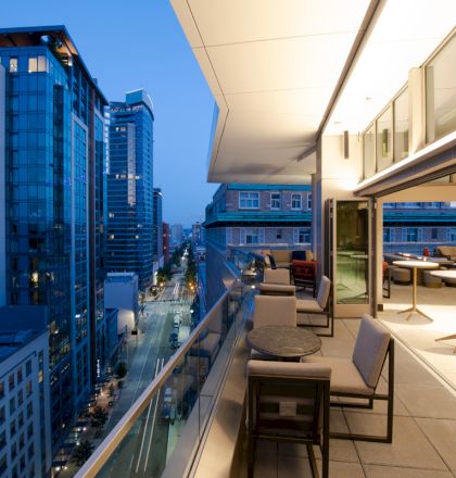 The image shows a modern cityscape view at dusk from a balcony with tables and chairs, overlooking tall buildings and a street below.