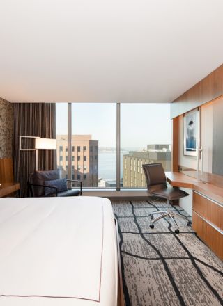 A modern hotel room with a large bed, desk, and chair. There's a flat-screen TV on the wall and a window with a city view.