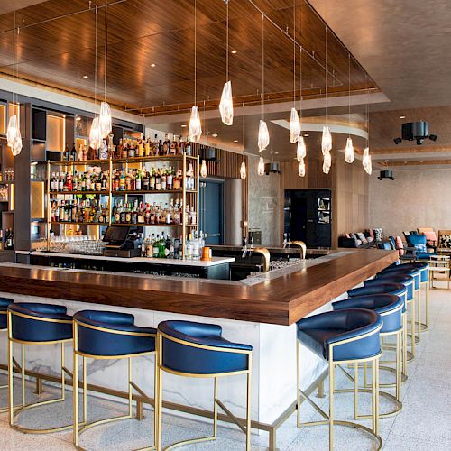 A modern bar with a central counter, blue chairs, hanging lights, and a well-stocked liquor shelf, surrounded by a cozy seating area.