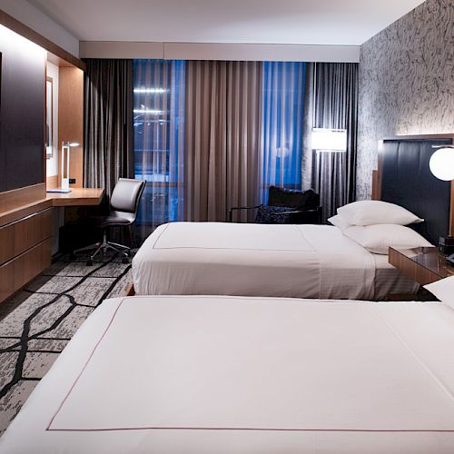 The image shows a modern hotel room with two beds, a TV, a desk, and decorative lighting, all in a sleek, contemporary design.