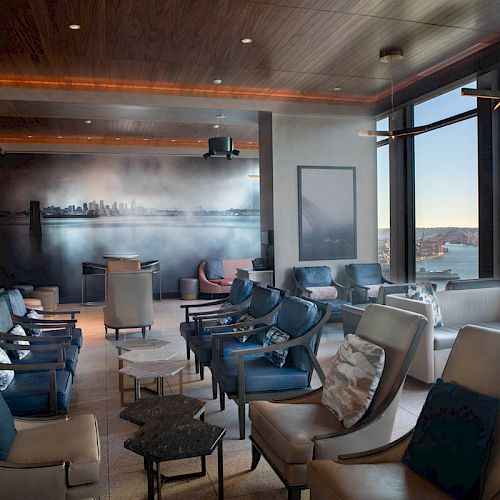 A modern lounge with comfortable seating, large windows viewing a cityscape and waterfront, and a mural on the back wall.